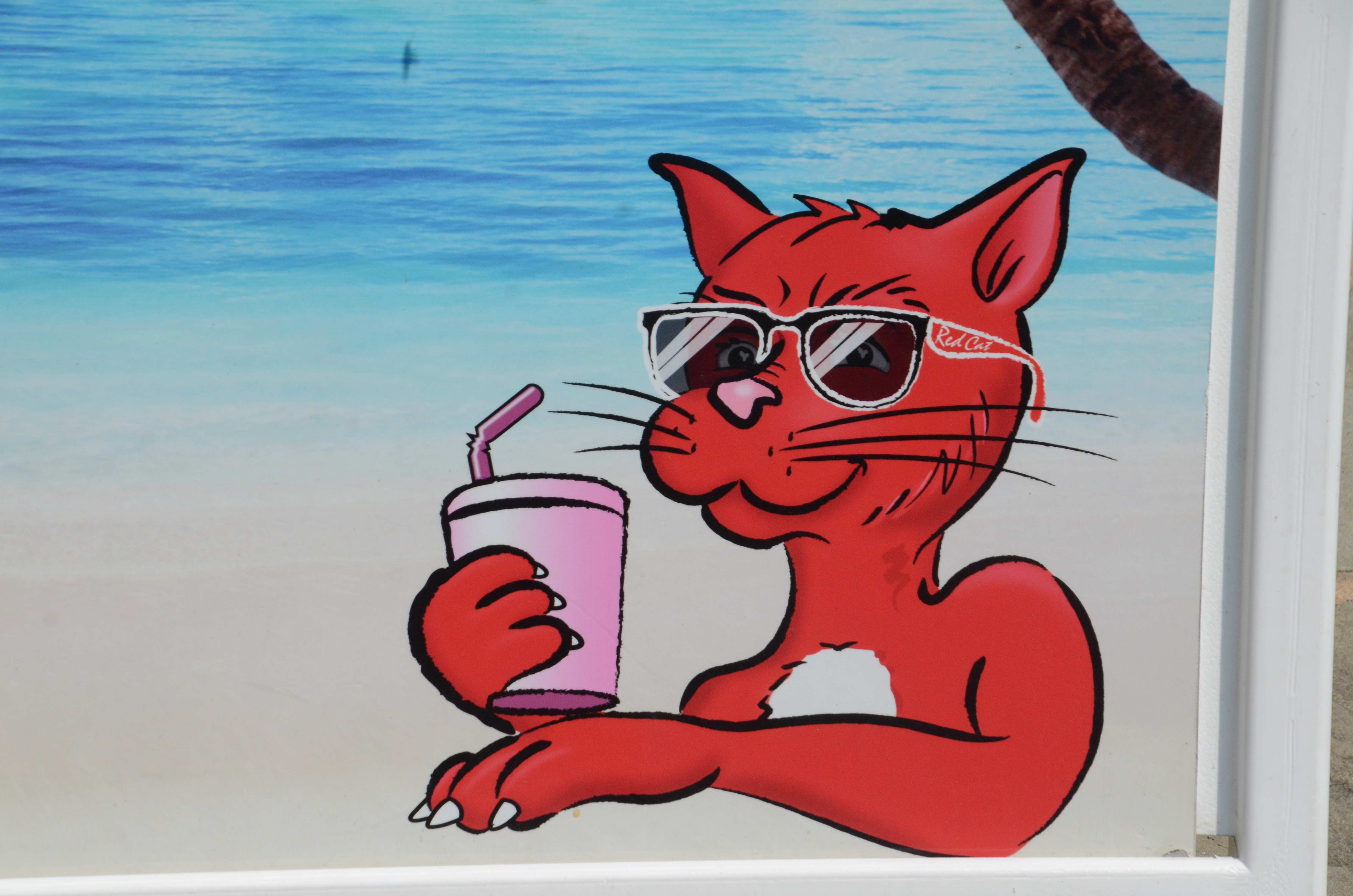 A picture of an advertisement for Red Cat wine. It features a cartoon cat, painted red, wearing sun glasses and enjoying a drink we can assume is alcoholic.