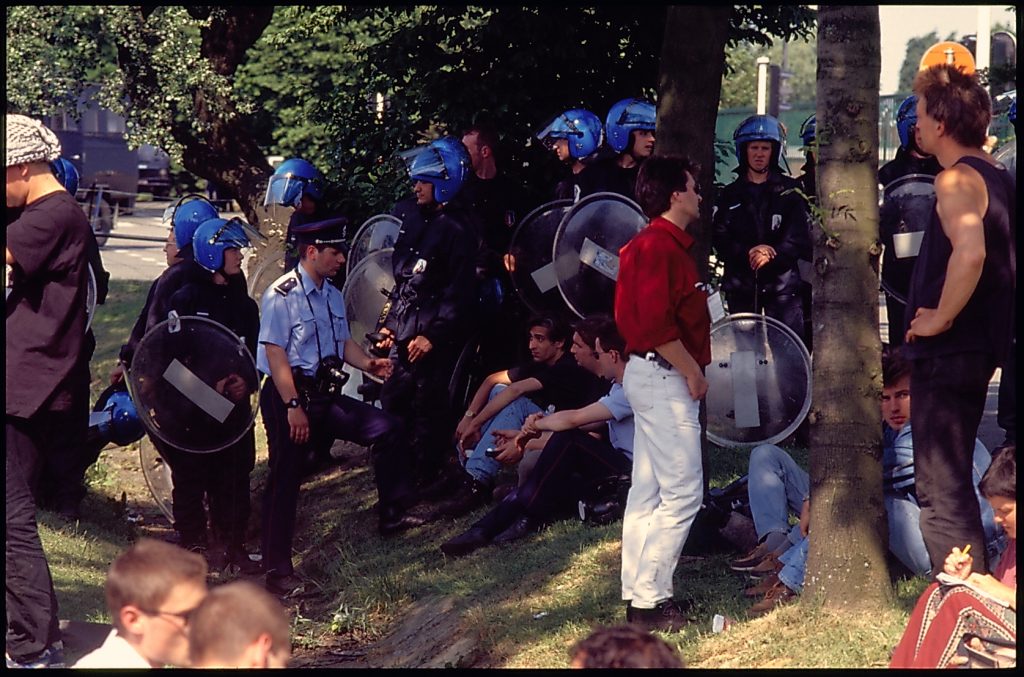 Riot police, and other officers in the shade talking with protesters.