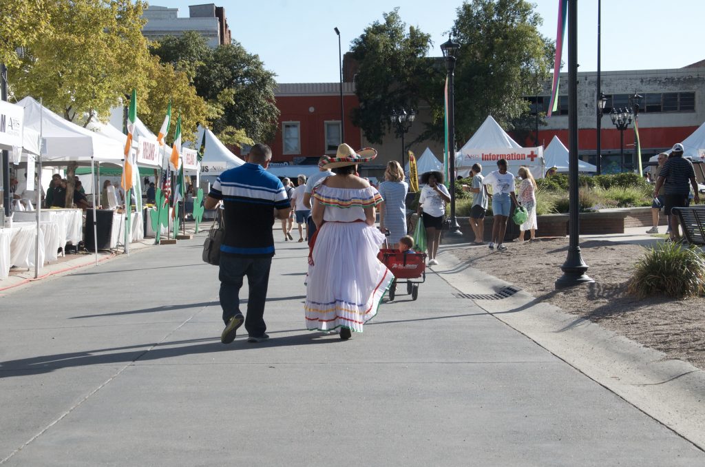 Two people walking away from the camera as the sun is setting. A few other people milling about.  The woman is wearing a traditional Mexican dress and hat.