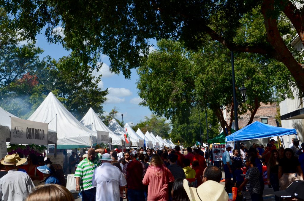 A crowd of people walking passed a series of white tents labeled with the names of countries like Sudan and Lebanon, where they are selling ethnic foods from those countries. 