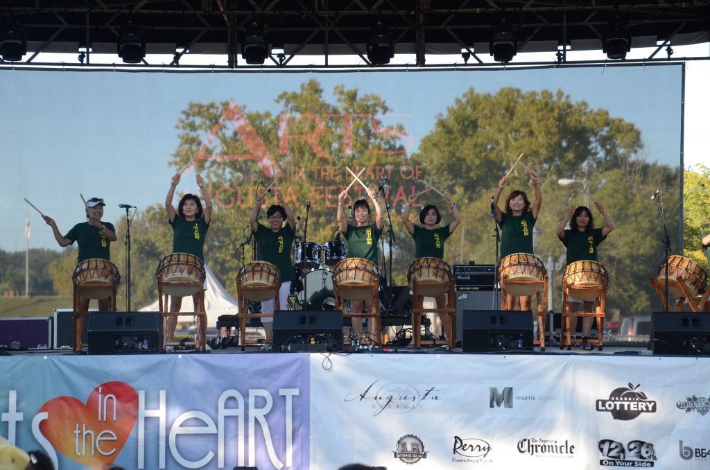 Image shows a collection of women of Chinese decent with green t-shirts with drum sticks raised as they perform on a large outdoor stage.