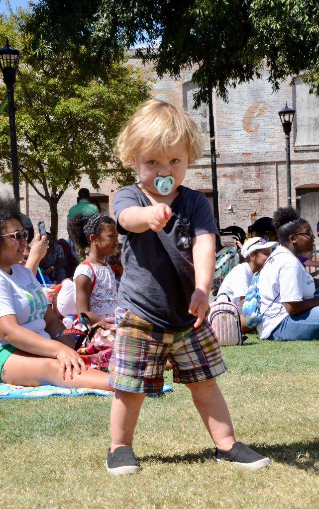 A little boy dancing in the grass, with a pacifier in his mouth, defiant look on his face, pointing at the camera.