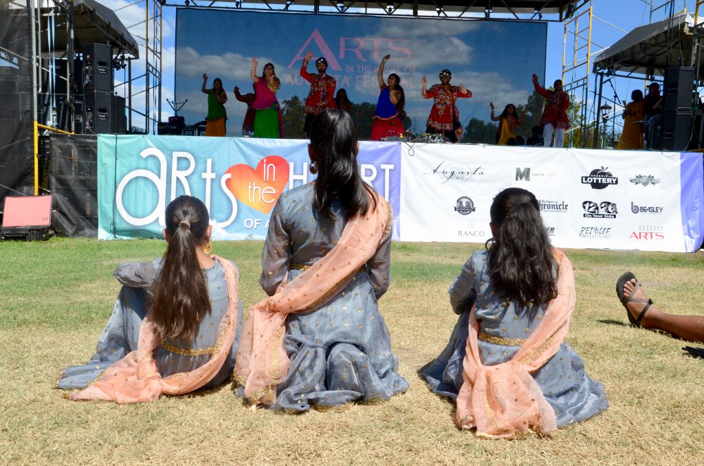 The girls who were just dancing, still dressed from their performance, are now kneeling on the green watching some older kids dance.