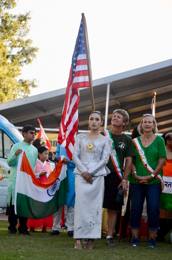 A woman of east asian decent, wearing a traditional dress, stands in front of a man wearing a sash from Ireland holding an American flag, while a boy of Indian decent stands next to them holding an Indian flag. All prepared to walk when their turn comes.
