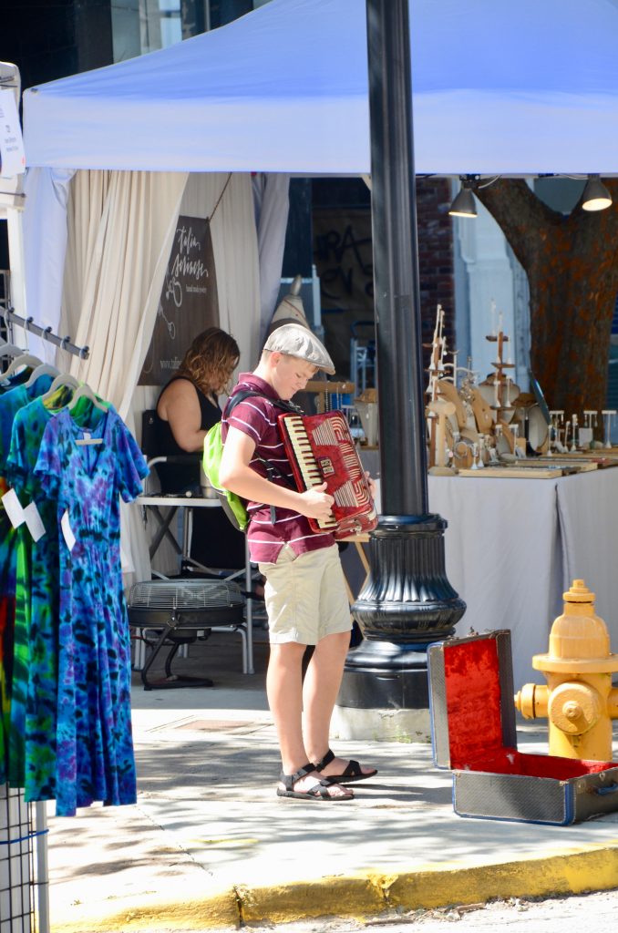 A boy standing on the side of the sidewalk playing an accordion, the case is open so people can leave tips.