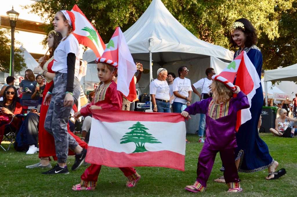 Small children and teenagers walking with the Lebanese flag. The small children traditional ethnic clothes, while the teenagers wear t-shirts and camo pants and have Lebanese flag head bands.