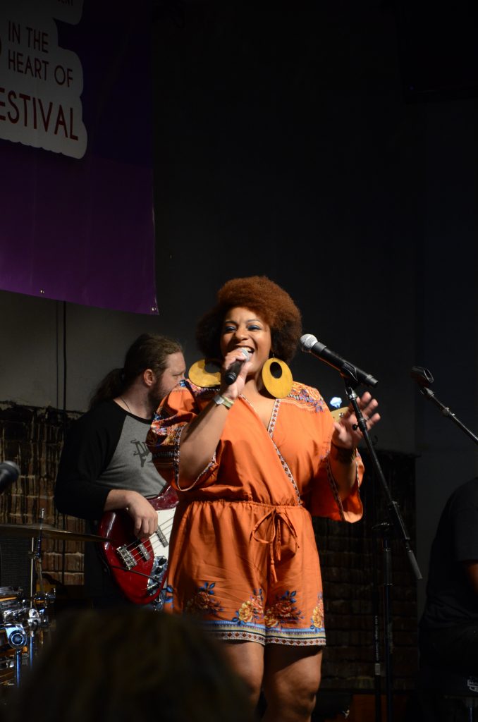 An African American woman singing on an indoor stage. She is clearly comfortable with the microphone and is smiling as she sings.