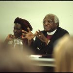 Desmond Tutu gesturing as he speaks in a crowded room.