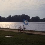 A bike with a peace dove flag flying in the breeze by a lake at sunset.