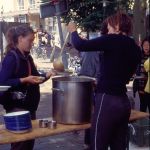 A woman serving soup from a large pot on a table setup in a town park.