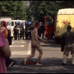 A naked man walking around in front of a line of police.