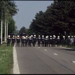 The backs of a line of police with black uniforms and white helmets.